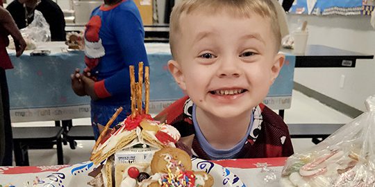A student smiles behind a gingerbread house