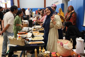 woman wearing a hijab flashes a peace sign with her fingers as she stands behind a table of various exotic foods