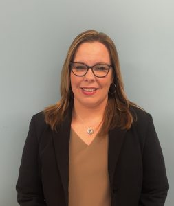 woman with shoulder length light brown hair, wearing glasses and a black jacket smiles for the camera