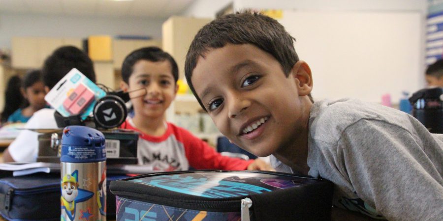 one student leaning over his lunchbox smiling and another student sitting tall smiling behind him